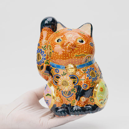 A gloved hand holds the copper-orange Kutani lucky cat, showing its size, raised right paw, blue collar and colorful floral decoration.