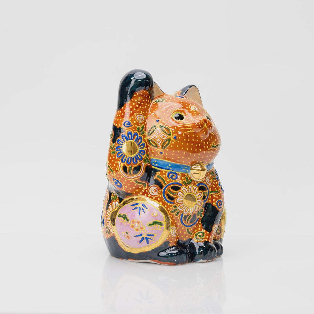 Three-quarter view of the Kutani lucky cat showing its raised paw, pink side panel, black patches and gold-tone floral centers.