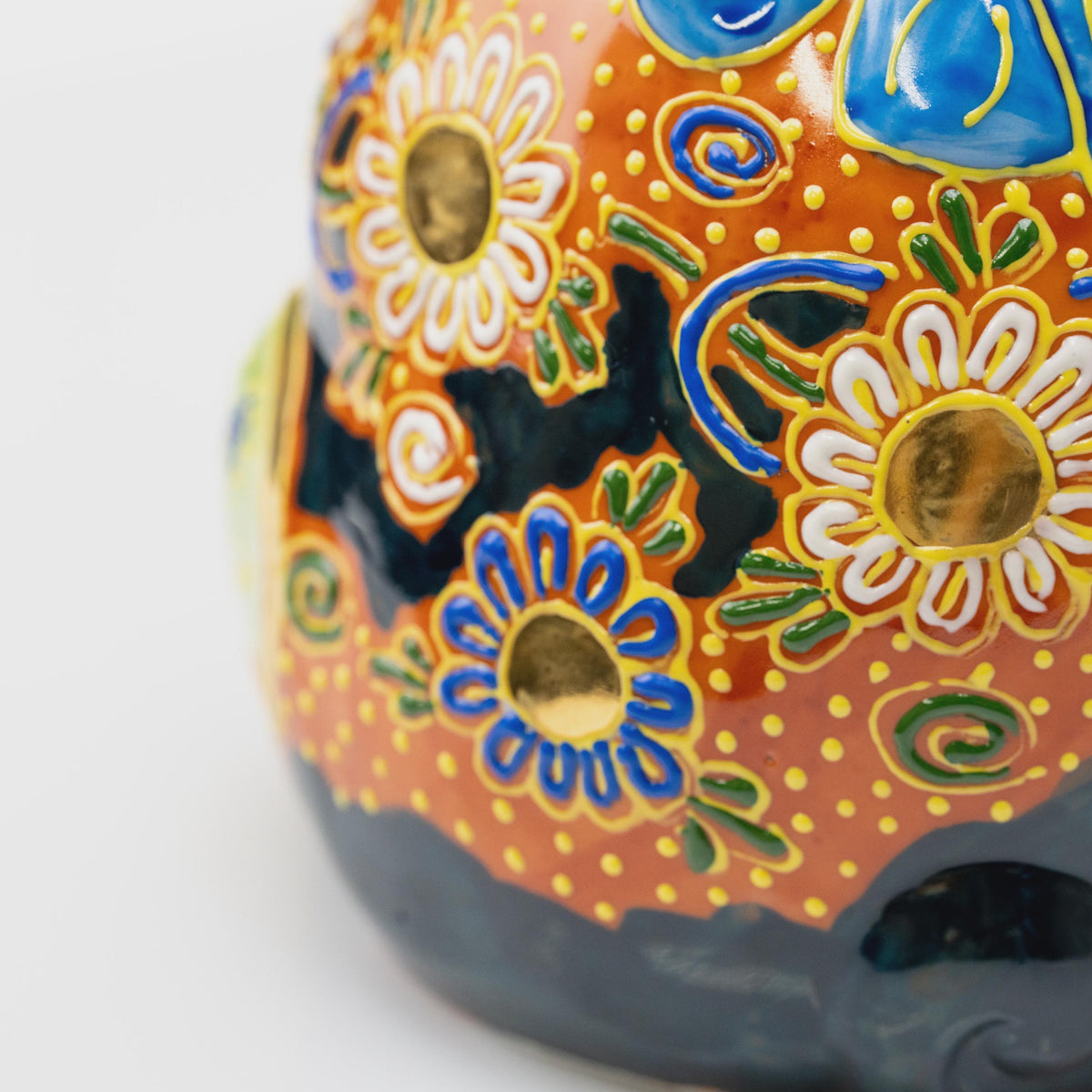 Close-up of blue and white floral decoration with gold-tone centers, green leaves and raised yellow dots on copper-orange glaze.