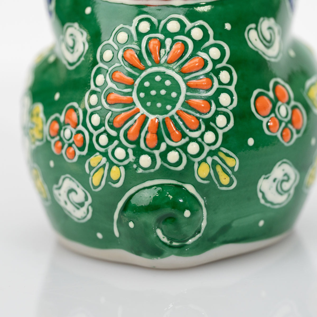 Close-up of the orange flower, white dots, and curled tail on the back of the green praying cat.