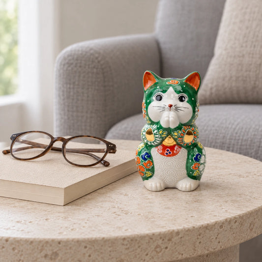 Green floral Kutani praying cat on a pale stone side table beside a book and reading glasses.