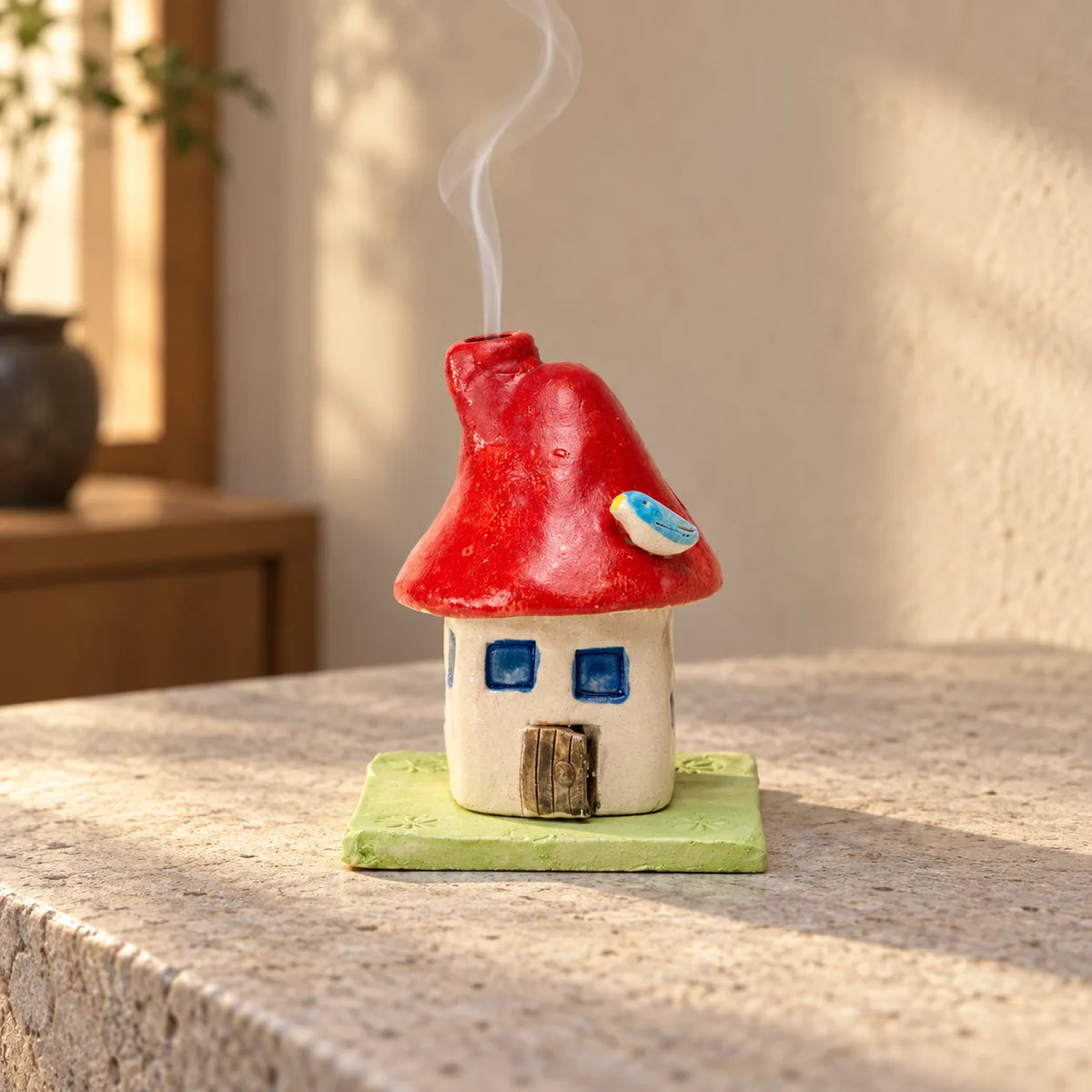 Red ceramic mushroom-house incense burner on its green tile with a thin wisp of smoke rising from the chimney