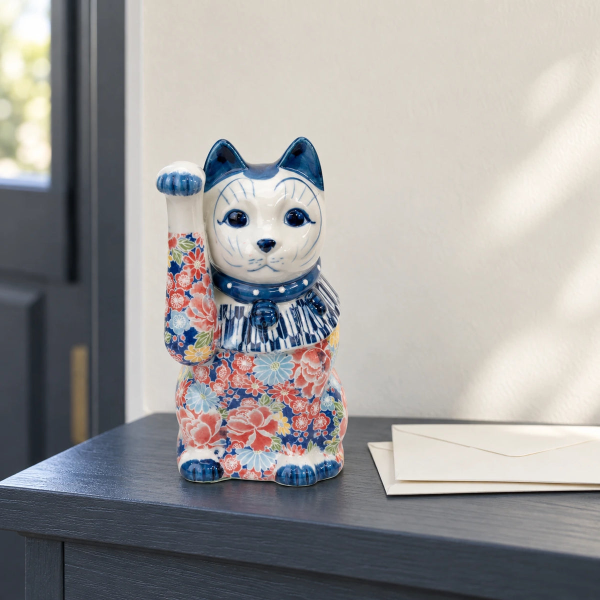 Maneki-neko Seto bleu à décor floral, patte droite levée, sur une tablette d’entrée gris anthracite à côté d’enveloppes ivoire