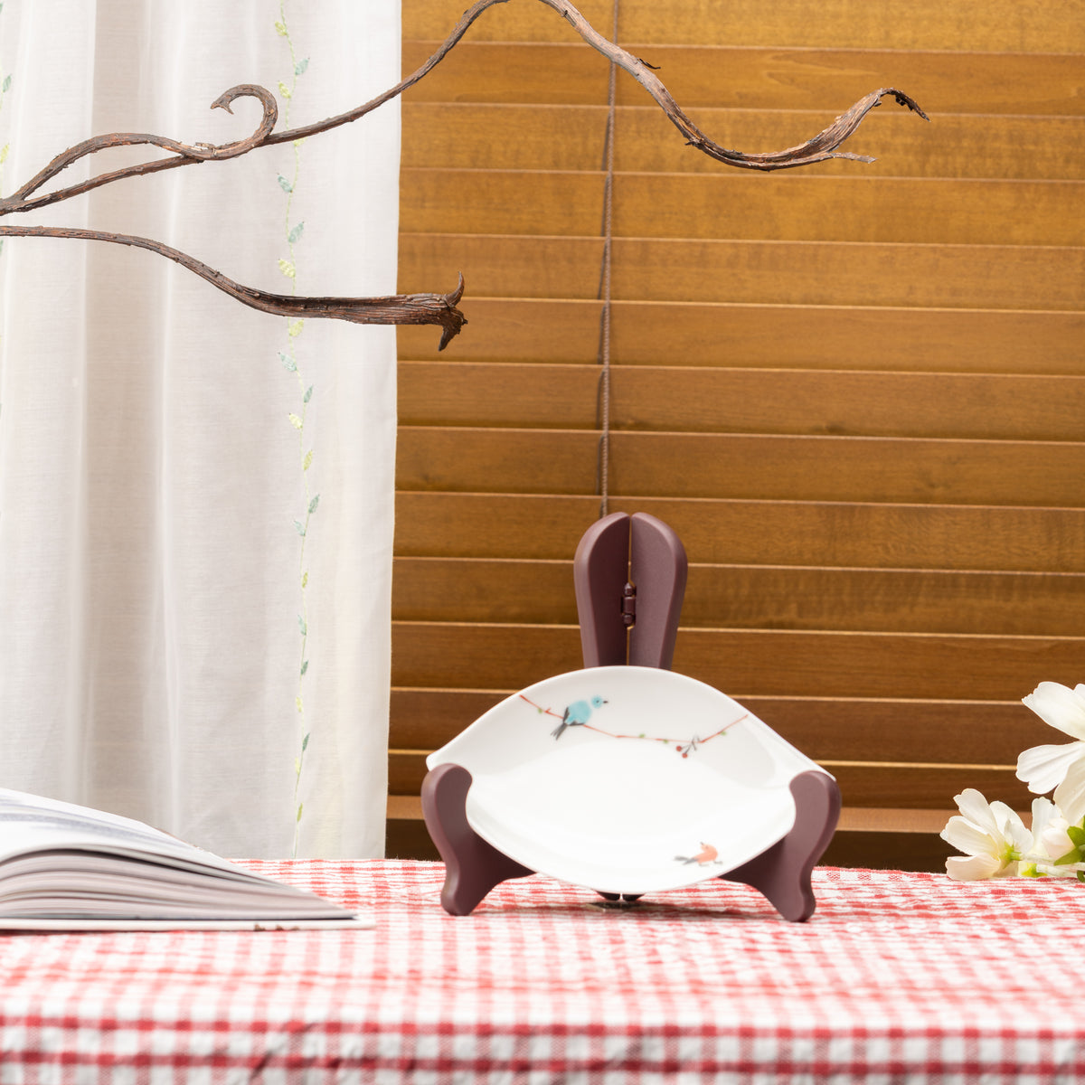 White Tsugumi bird leaf plate displayed on a stand on red gingham beside an open book and flowers.