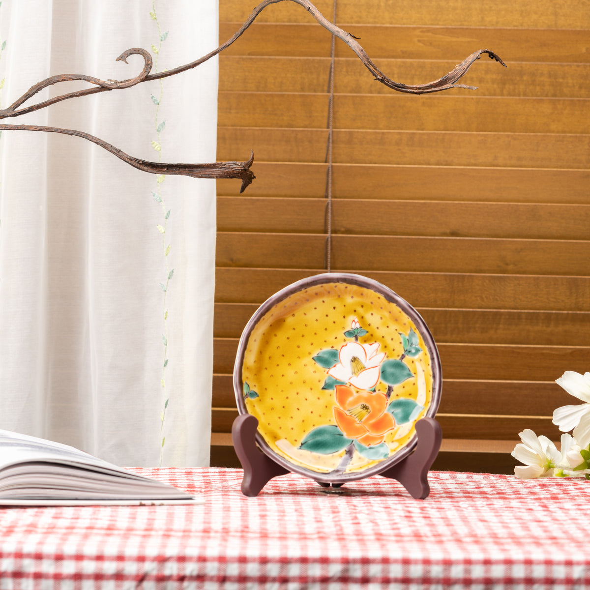 Yellow camellia plate on a stand beside an open book, on a red gingham tablecloth by a window.