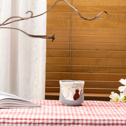 Hidamari cat yunomi displayed on a red gingham tablecloth beside an open book and window blinds.