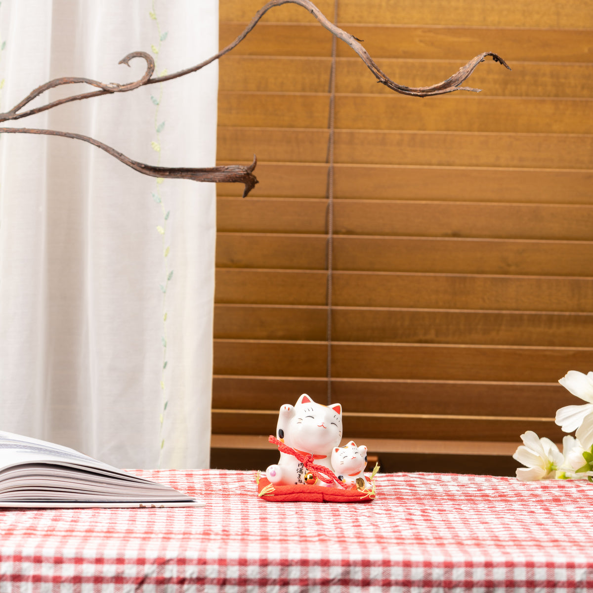 Parent-and-kitten lucky cat on a red cushion, displayed on a gingham tablecloth beside an open book.