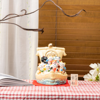 Seven lucky cats treasure ship displayed on red gingham beside an open book, flowers and a small sign.