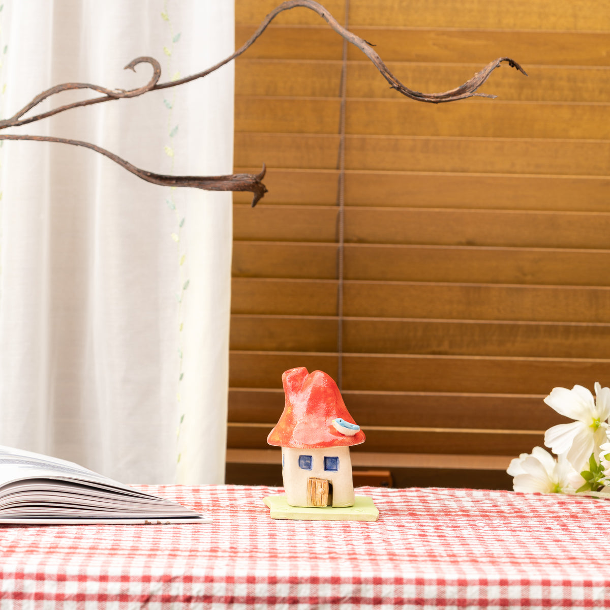Red-roofed mushroom house incense burner displayed on a red gingham tablecloth by a window.