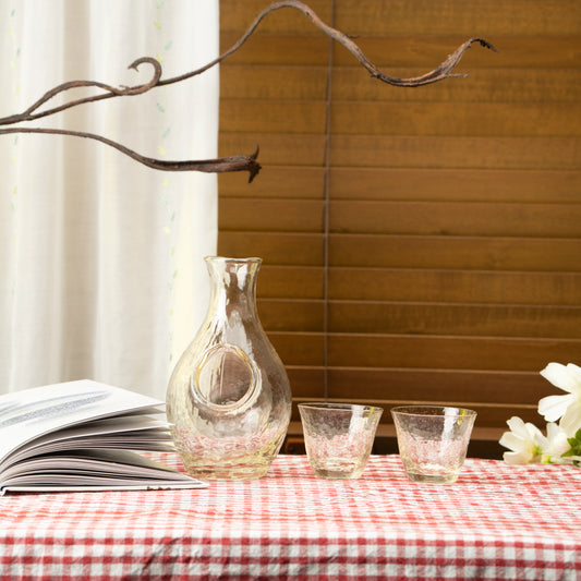 Amber glass sake carafe and two cups on a red checked tablecloth beside a book.