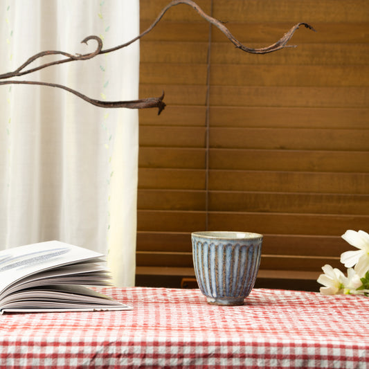 Blue-gray ribbed yunomi on a red checked tablecloth beside a book and white flowers.