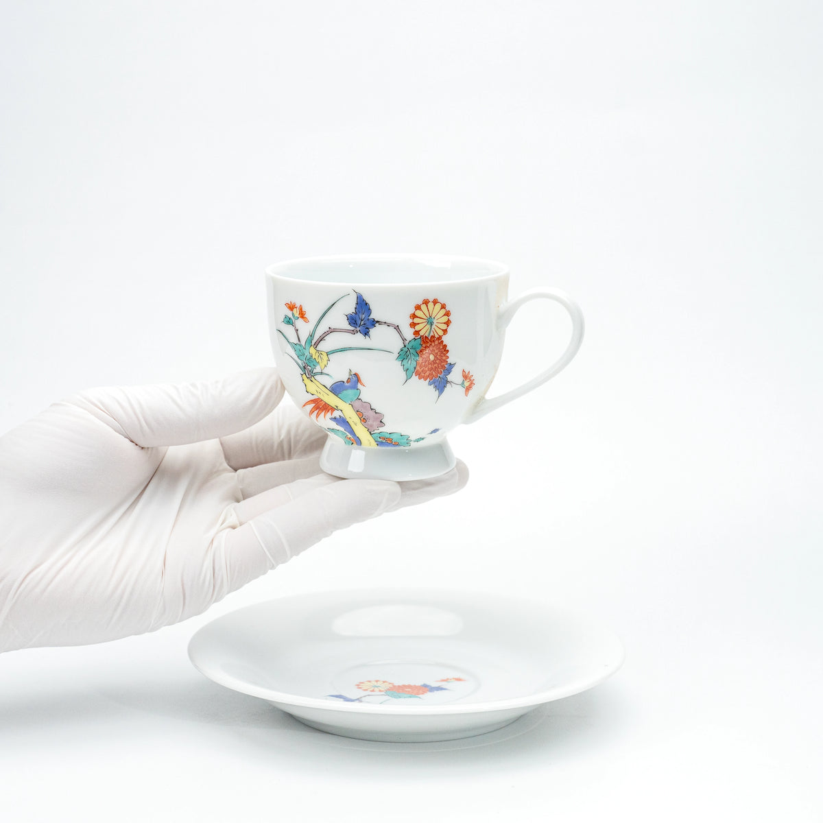 Porcelain cup with chrysanthemums and birds held in a gloved hand.