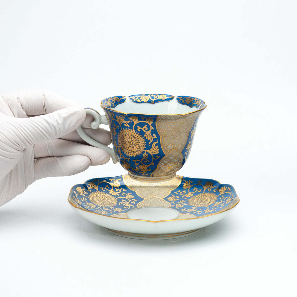 Blue and gold cup held above its saucer in a gloved hand.