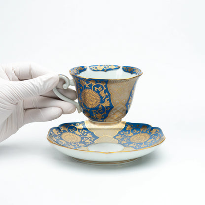 Blue and gold cup held above its saucer in a gloved hand.