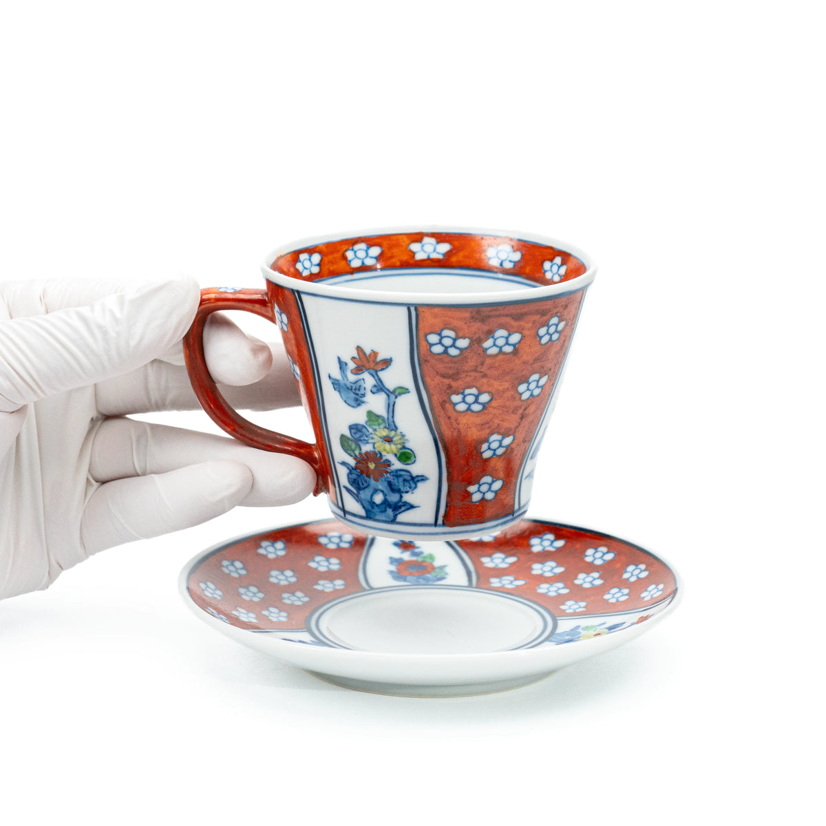 Red floral cup held above its matching saucer in a gloved hand.