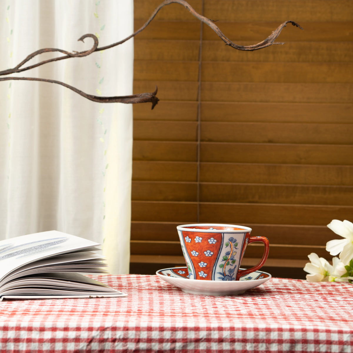 Red floral cup and saucer on a red checked tablecloth beside a book.