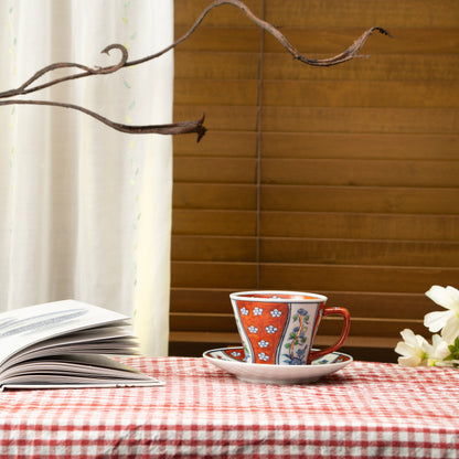 Red floral cup and saucer on a red checked tablecloth beside a book.