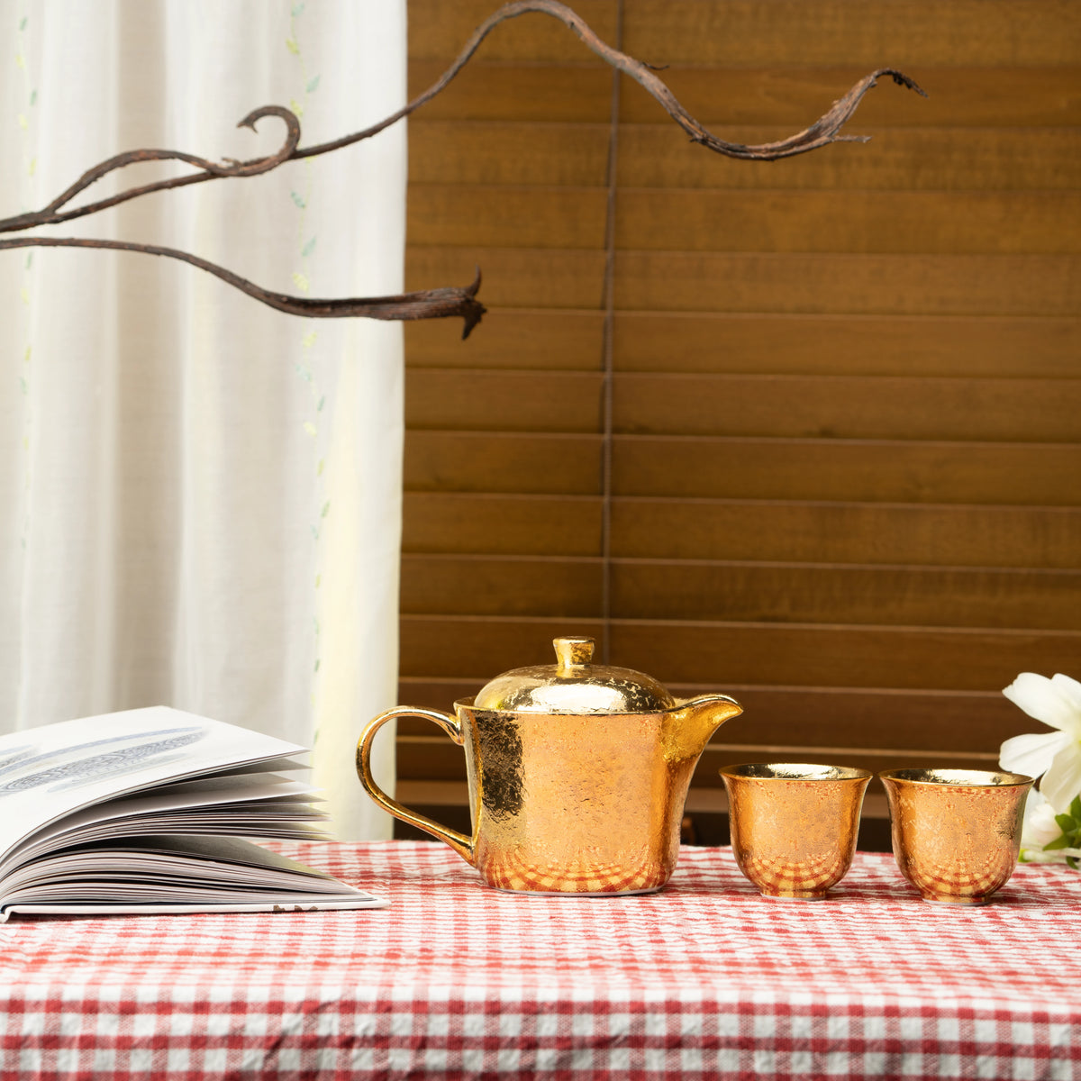 Gold tea set on a red checked tablecloth beside a book and flowers.