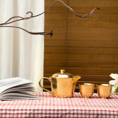 Gold tea set on a red checked tablecloth beside a book and flowers.