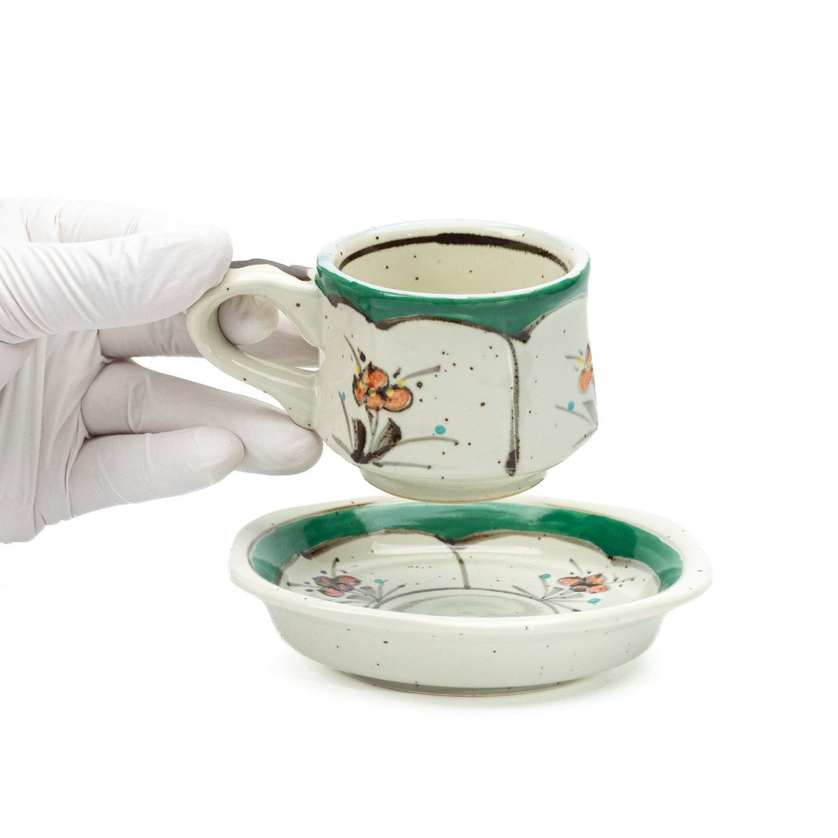 Small green floral cup held above its saucer in a gloved hand.