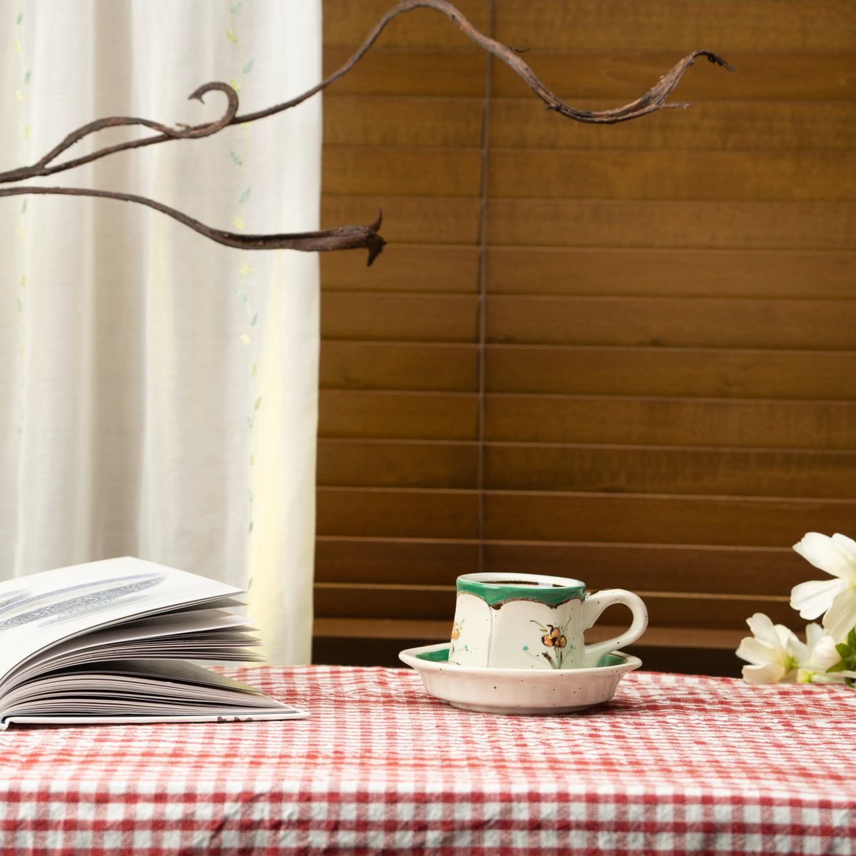 Green floral demitasse cup and saucer on a red checked tablecloth beside a book.