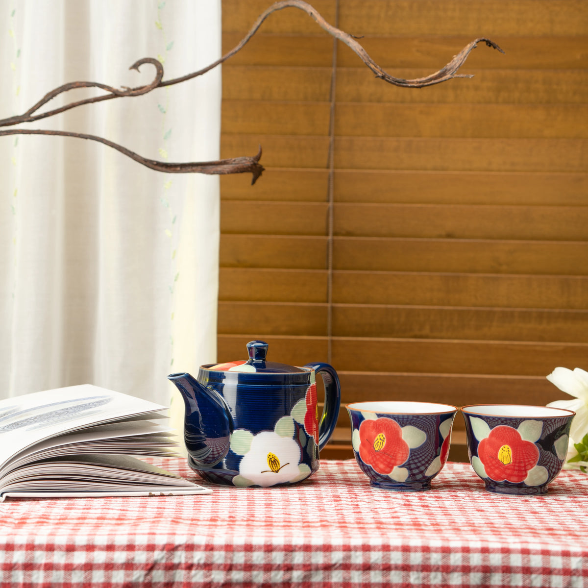 Blue camellia tea set on a red checked tablecloth beside a book and flowers.