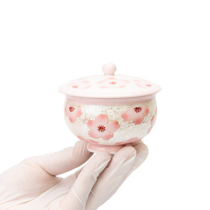 Pink cherry blossom lidded teacup held in a gloved hand.