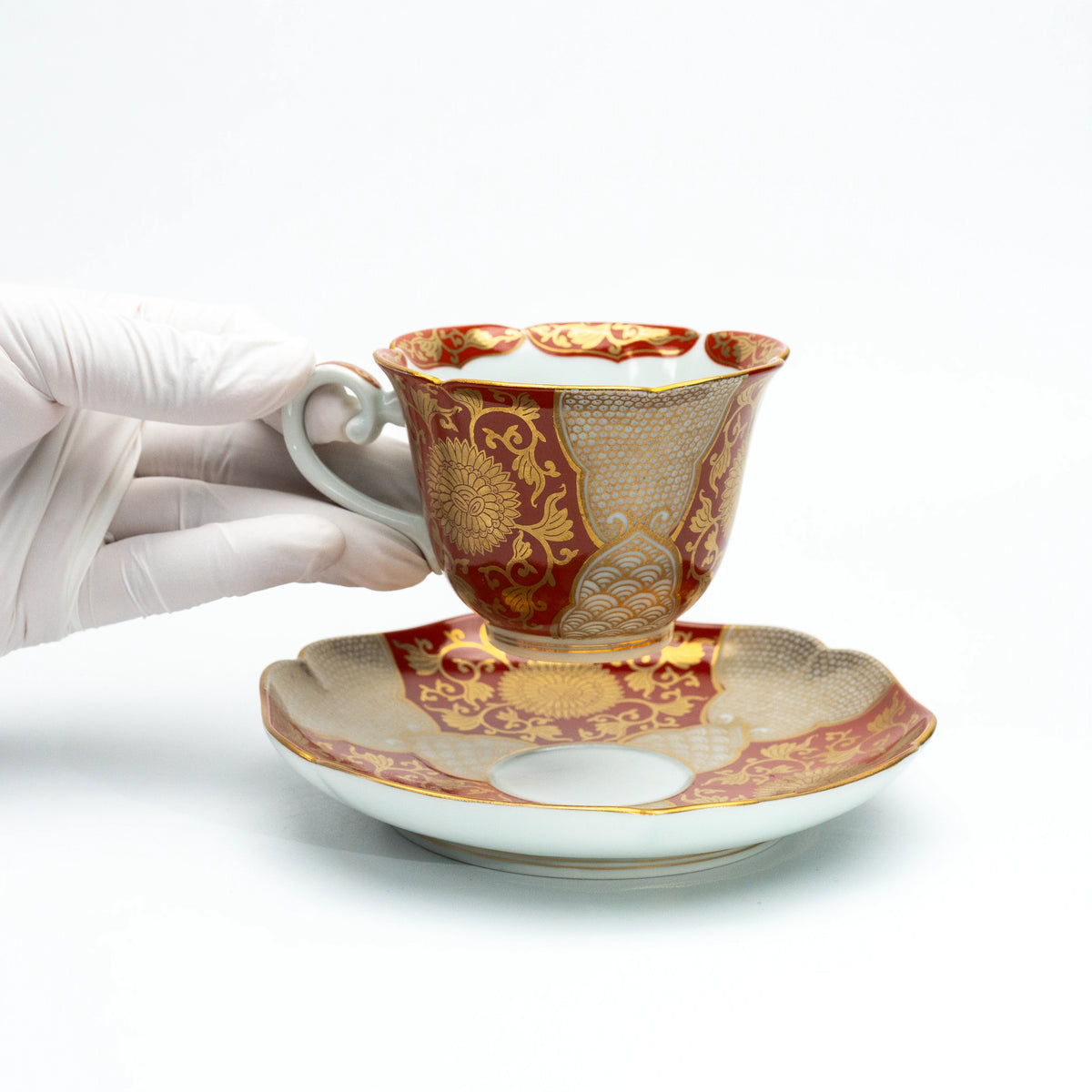 Red and gold cup held above its matching saucer in a gloved hand.