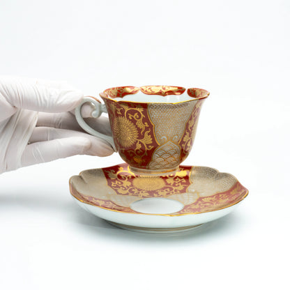 Red and gold cup held above its matching saucer in a gloved hand.