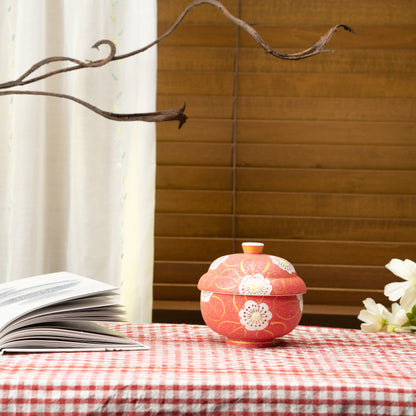 Red floral lidded bowl on a red checked tablecloth beside a book.