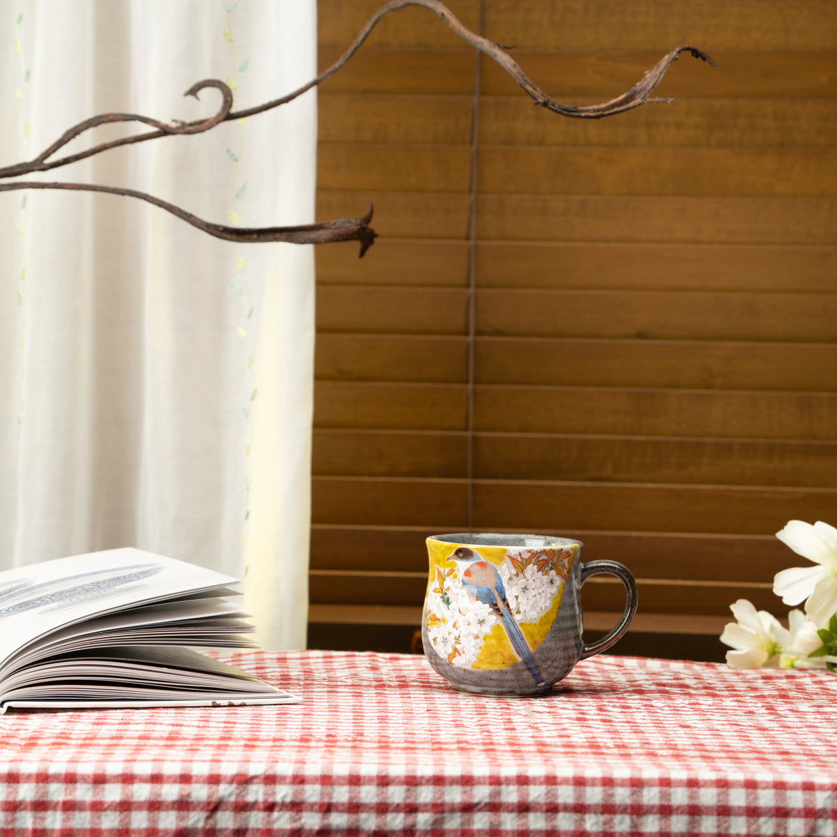 Yellow bird and cherry blossom Kutani mug on a red checked tablecloth beside a book.