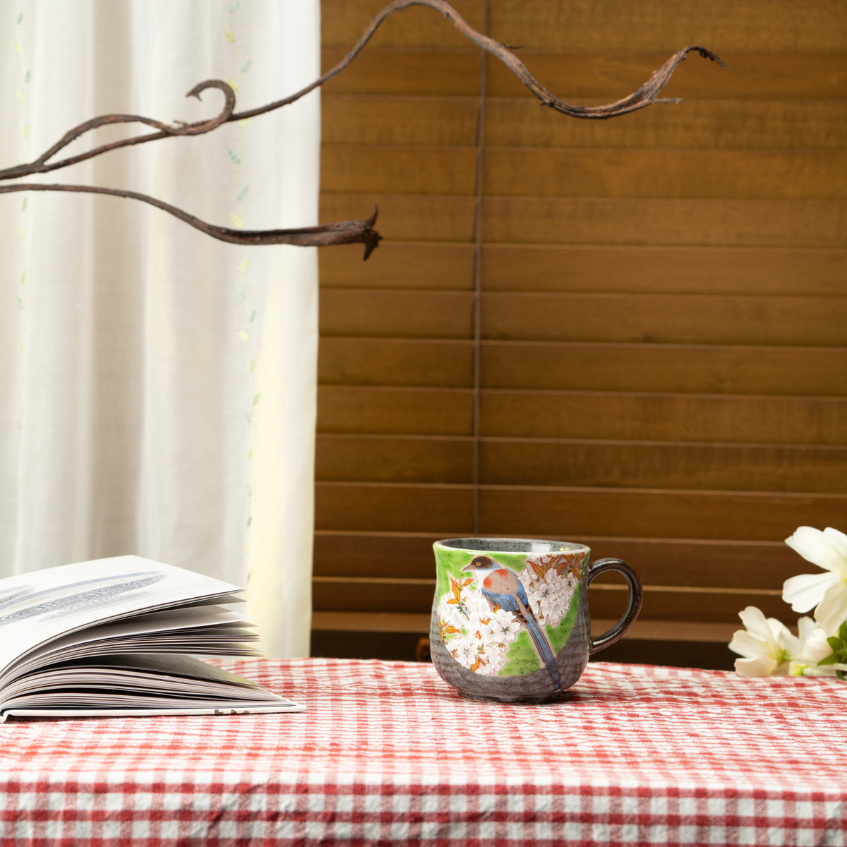 Green bird and cherry blossom Kutani mug on a red checked tablecloth beside a book.