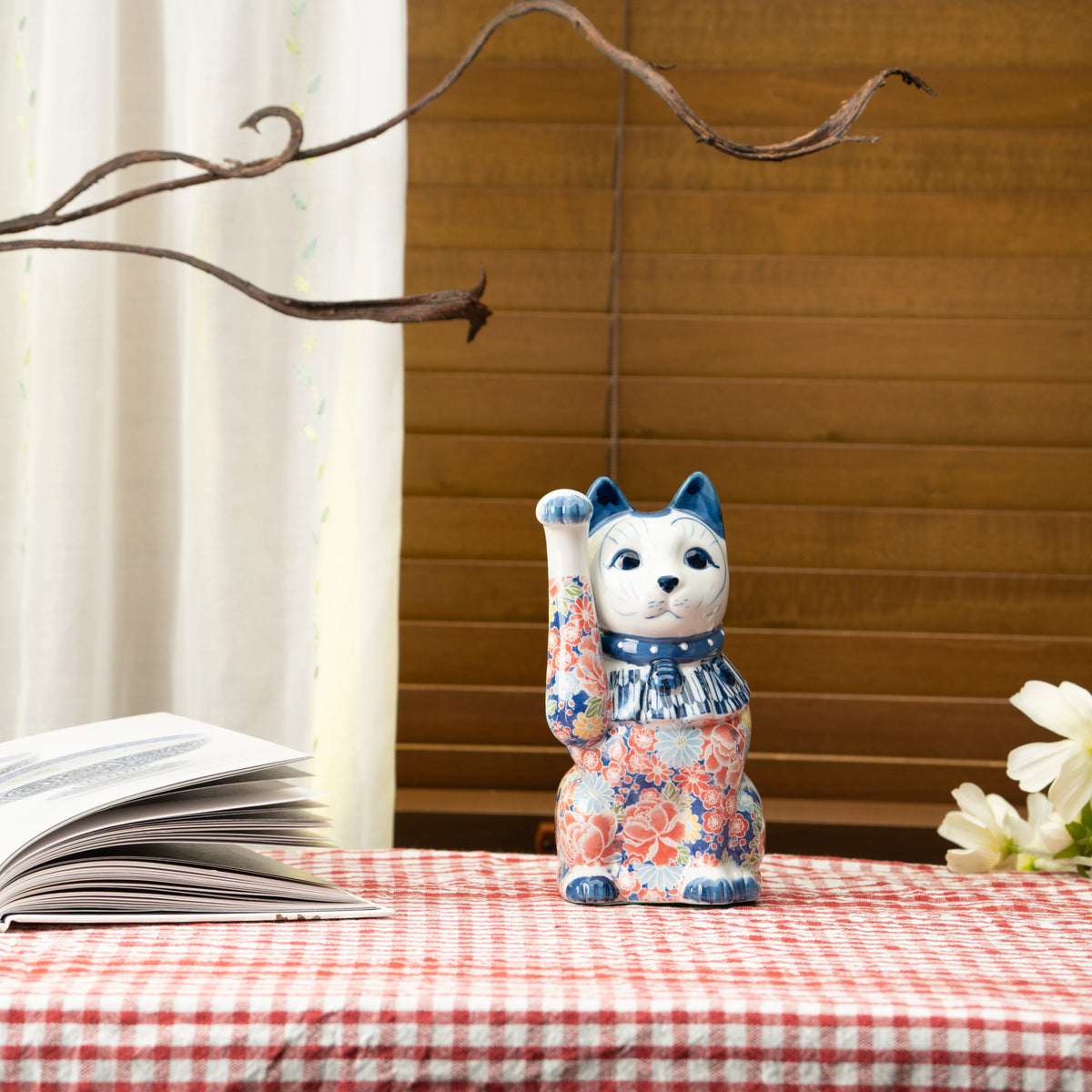 Blue floral lucky cat on a red checked tablecloth beside a book.