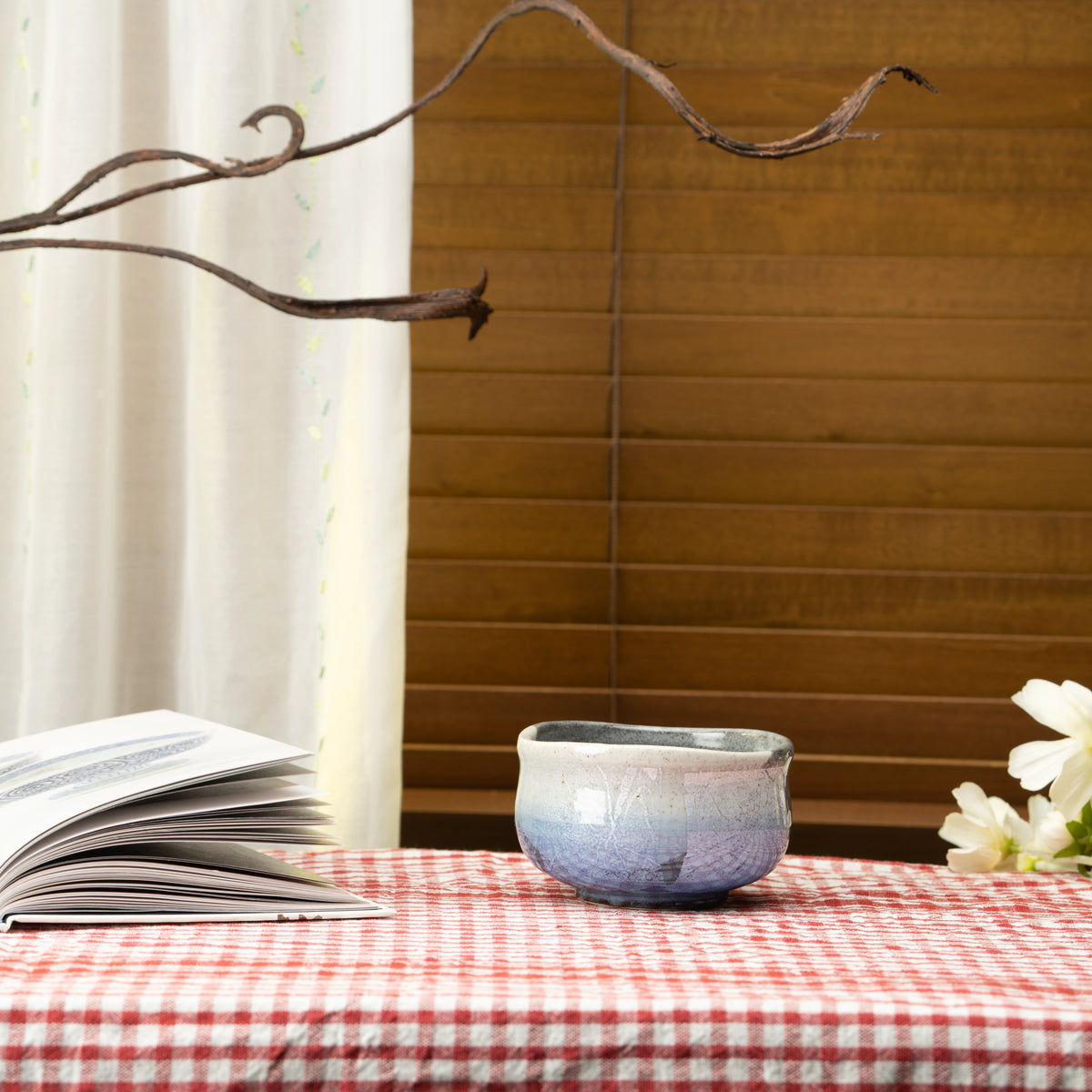 Blue ginsai matcha bowl on a red checked tablecloth beside a book and white flowers.