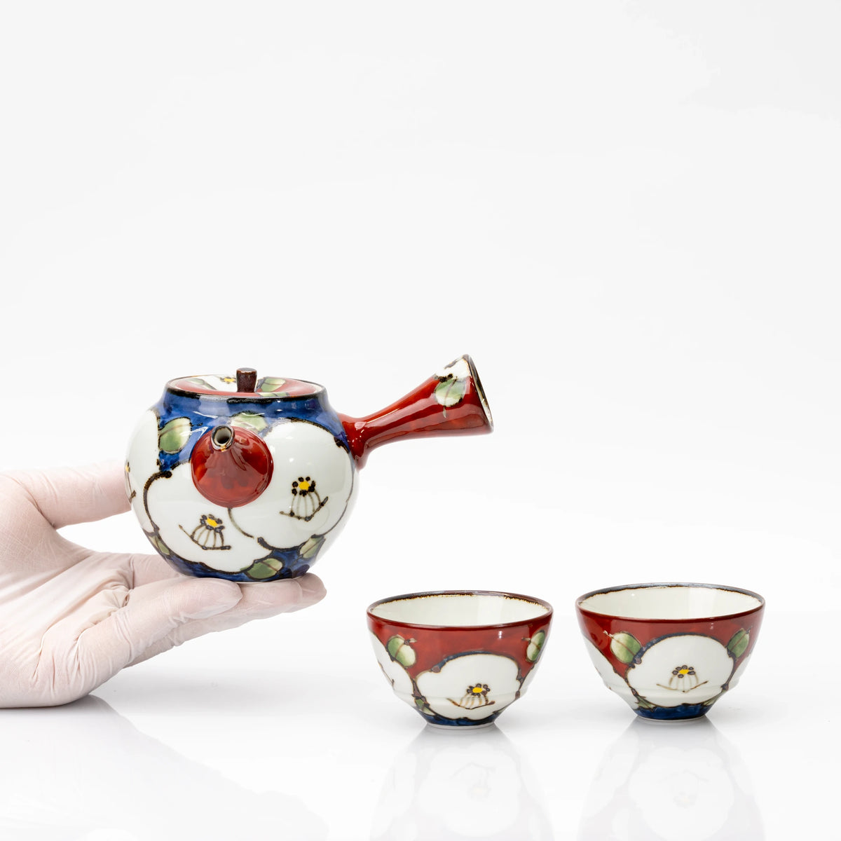 Gloved hand holding the camellia teapot beside two matching red cups, showing the compact size of the set.