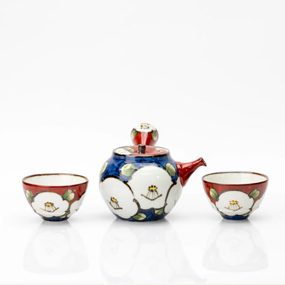 Front view of a blue porcelain camellia teapot between two red cups, with white flowers and pale green leaves.