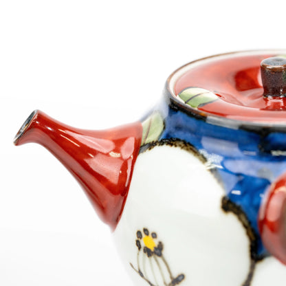 Close-up of the teapot’s red spout beside the blue body, white camellia decoration and red lid.
