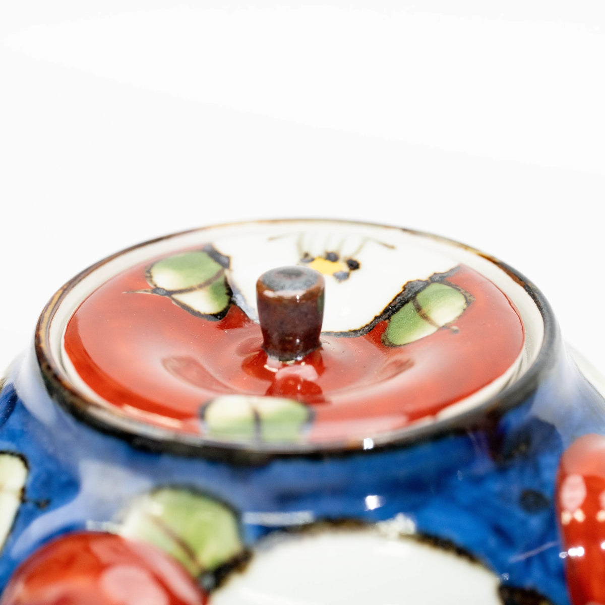 Close-up of the red teapot lid with a dark cylindrical knob, white camellia petals and green leaves.