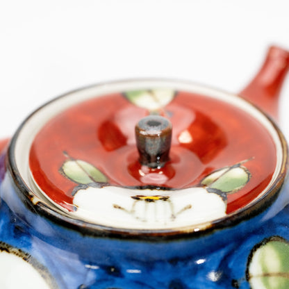 Angled close-up of the glossy red teapot lid and dark knob, with white camellia and green leaf decoration.