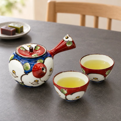 Blue camellia kyusu and two red cups of green tea on a gray dining table, with small sweets in the background.