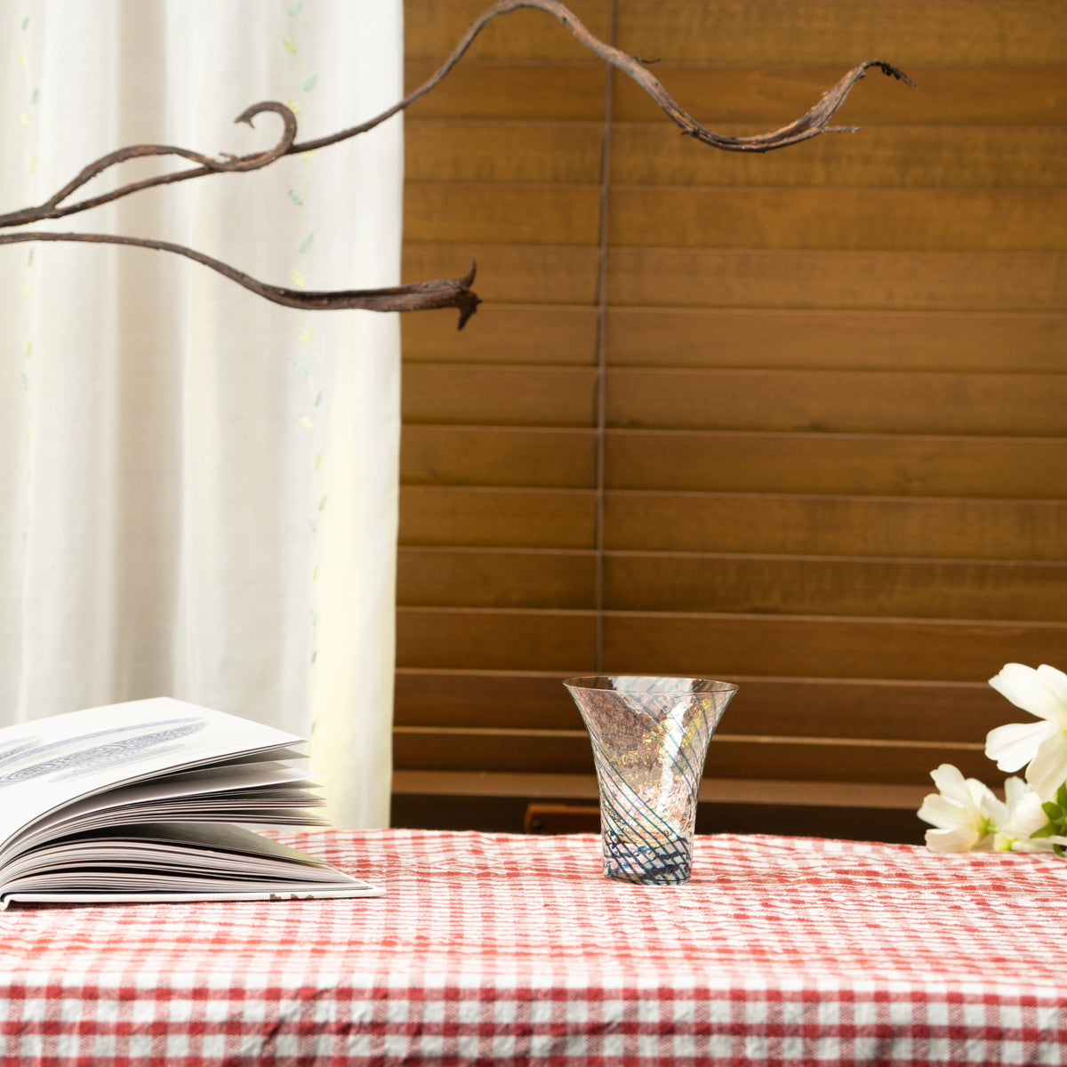 Blue and gold-colored Yachiyo glass sake cup on a red checked tablecloth beside a book.