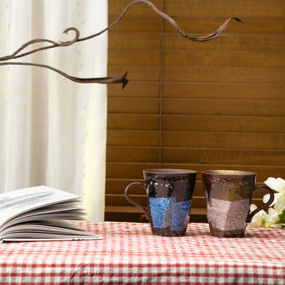 Blue and lilac Kutani mug pair on a red checked tablecloth beside a book and flowers.