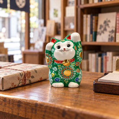 Petit chat porte-bonheur Kutani vert, les deux pattes levées, sur le comptoir d’une librairie à côté d’un livre emballé