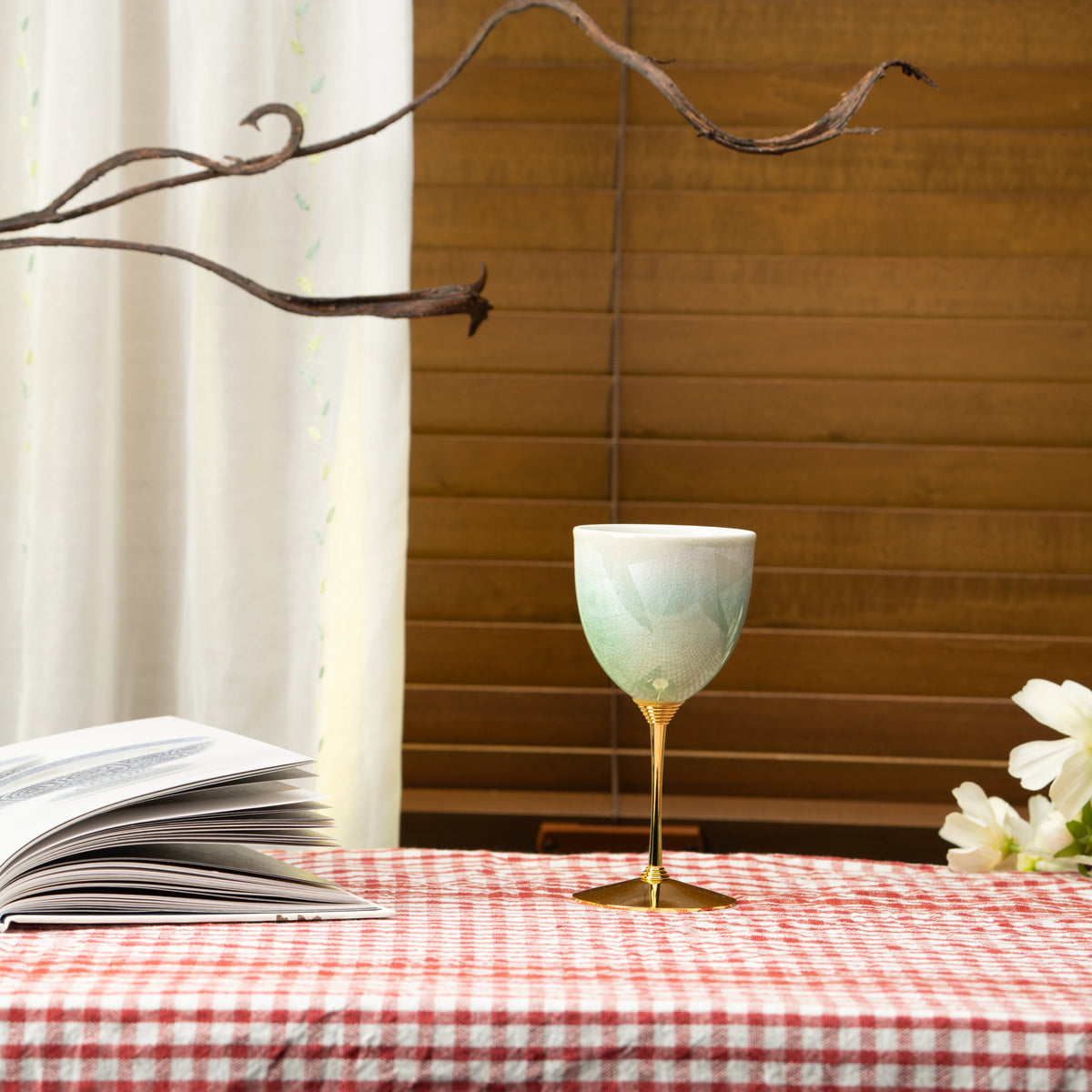 Green Kutani wine cup on a red checked tablecloth beside a book and white flowers.