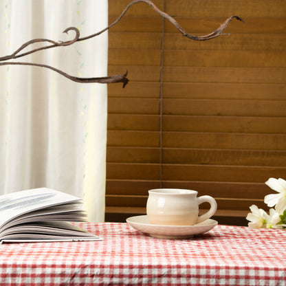 Cream and beige Hagi cup and saucer on a red checked tablecloth beside a book.