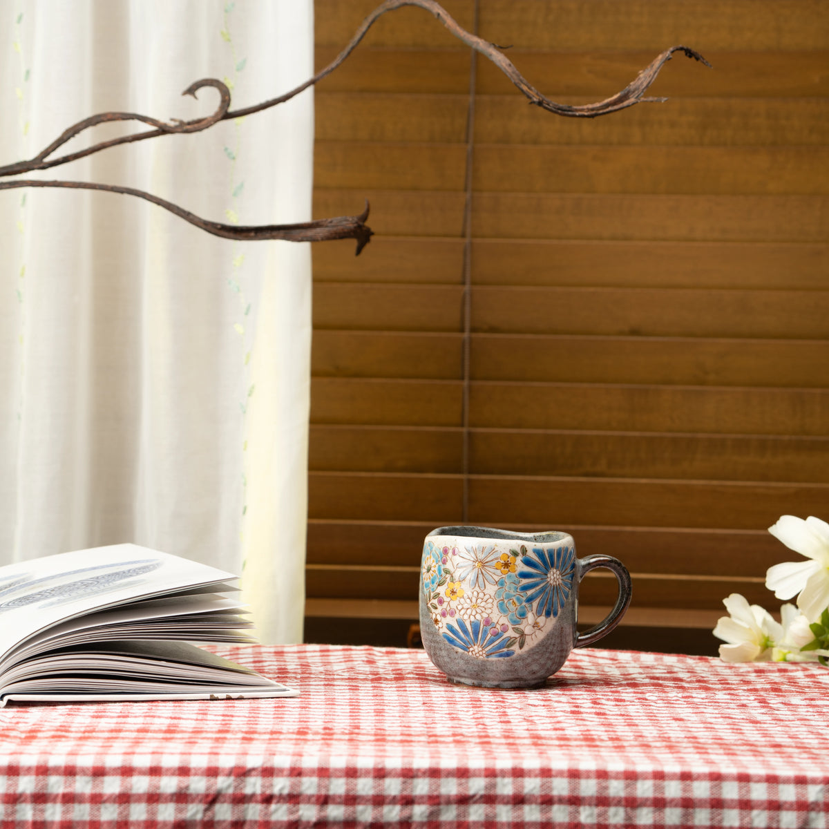Blue floral Kutani mug on a red checked tablecloth beside a book and white flowers.