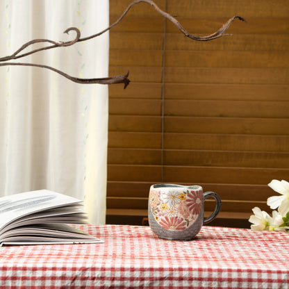 Red floral Kutani mug on a red checked tablecloth beside a book and white flowers.