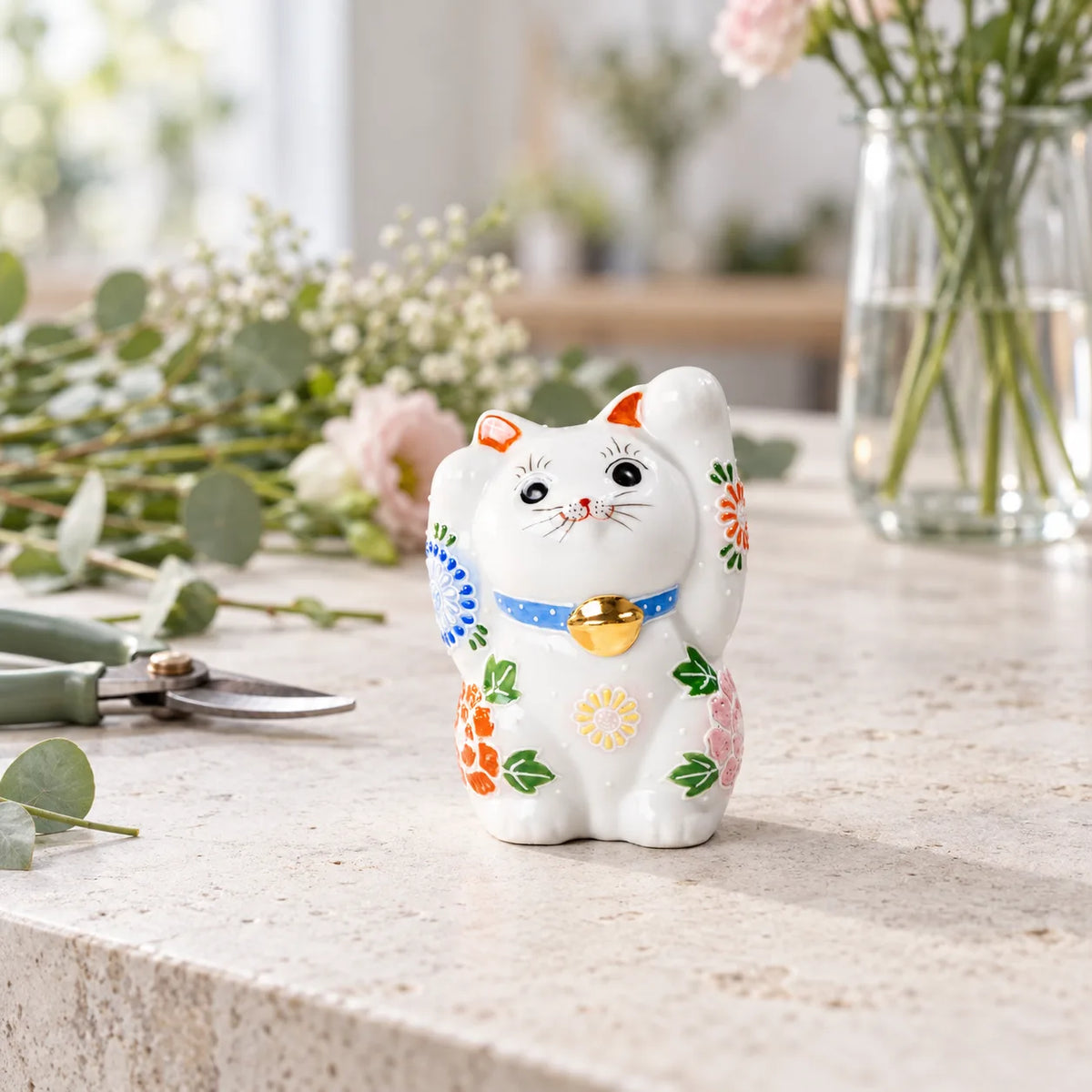 White Hanazono lucky cat with raised floral details on a florist worktable beside cut stems and pruning snips