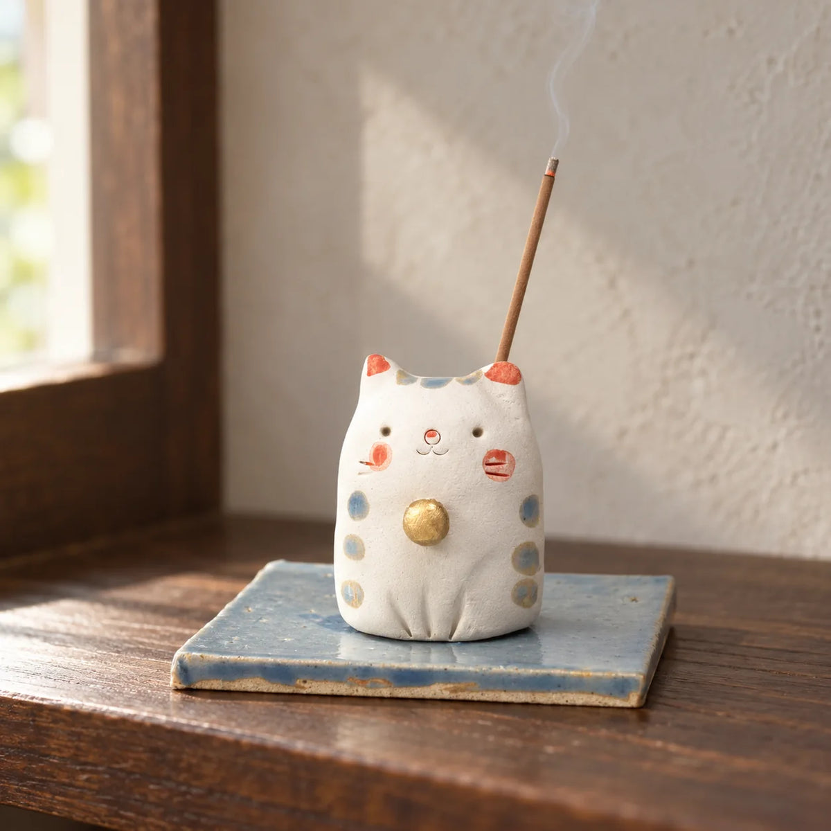 Handmade white cat incense holder on its blue ceramic dish with a short lit incense stick inserted at the back