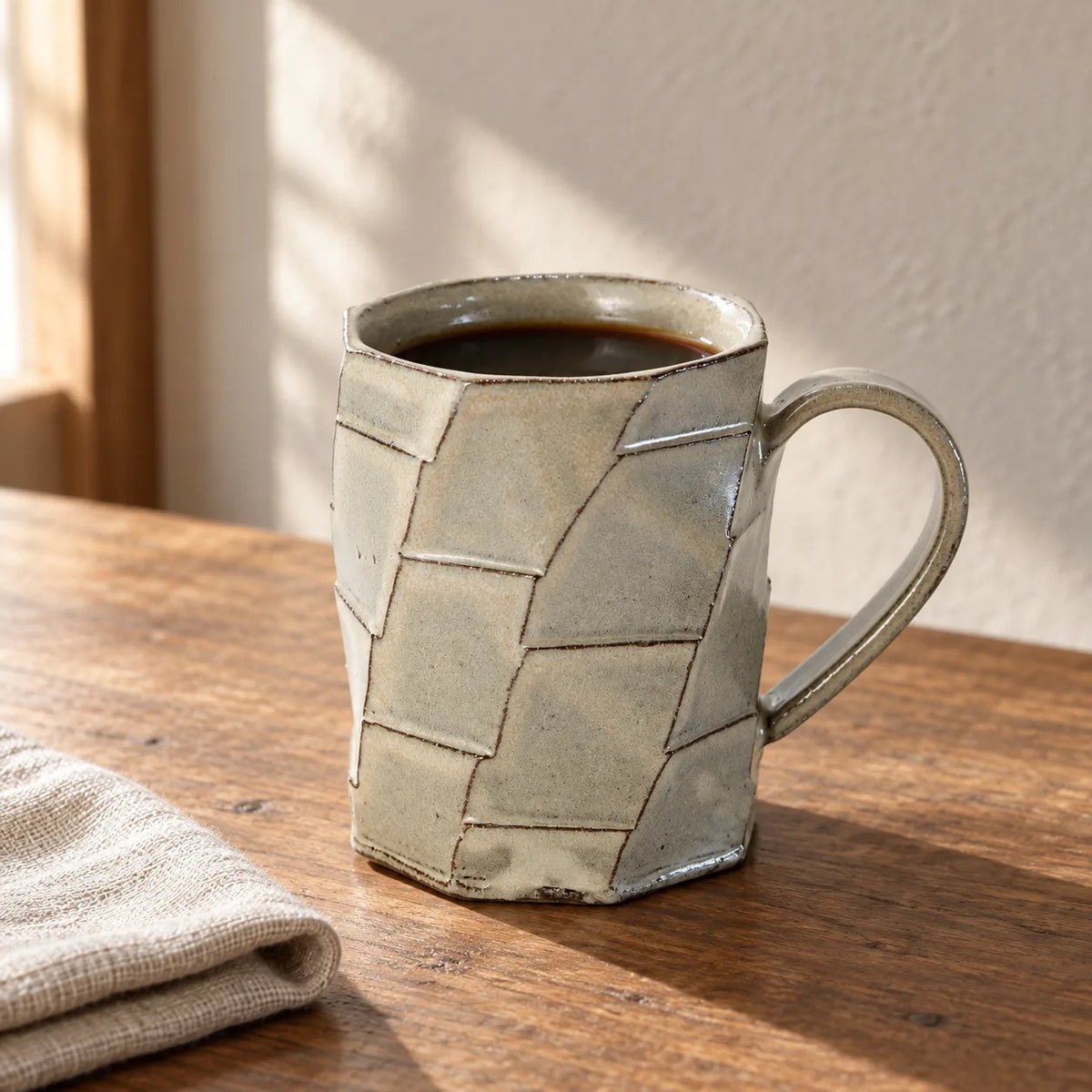 Faceted greige Shigaraki mug filled with black coffee on a wooden table beside folded linen.